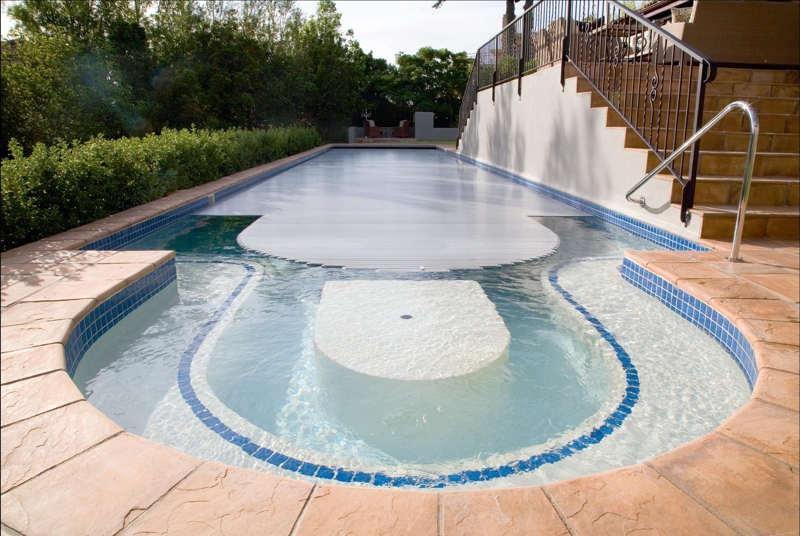 Powerplastic Pool Covers (021 703 5880). The owners of this home requested that the PowerPlastics Roldeck be able to cover the submerged cocktail table and seats contained within the Roman end of the pool, and that the slats conform to the curve of the pool. Featured here is the pool cover half open, showing the curve of the pool and the seating area and clearly demonstrating the fine engineering of the cover itself. When fully retracted, the cover withdraws into a concealed pit, leaving the pool completely open for the enjoyment of the owners.